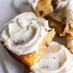 Two cinnamon rolls topped with cream cheese frosting on a plate. More cinnamon rolls in the background.