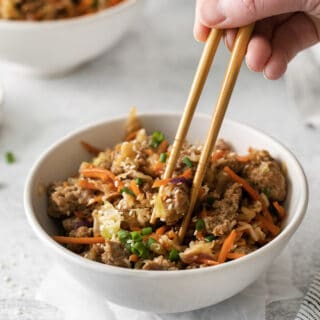 Photo of a hand holding a pair of chopsticks, and picking up a bite of Egg Roll in a Bowl from a white bowl. There is another white bowl in the background.