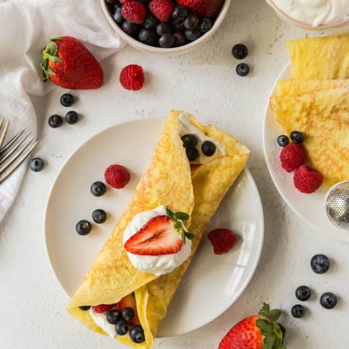 An almond flour crepe filled with keto whipped cream, blueberries, raspberries and strawberries. Its also topped with whipped cream and a strawberry. There are berries scattered around the countertop and a plate with more creapes.