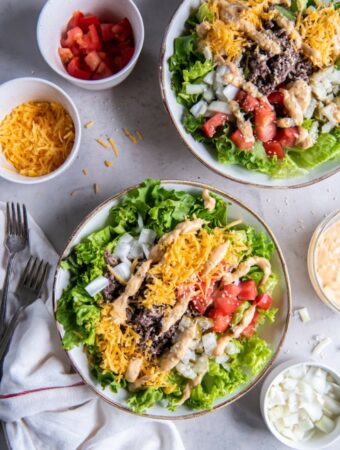Two plates of salad greens containing cooked ground beef, shredded cheddar cheese, diced red tomatoes, diced white onions, drizzled big mac sauce and a sprinkle of sesame seeds.