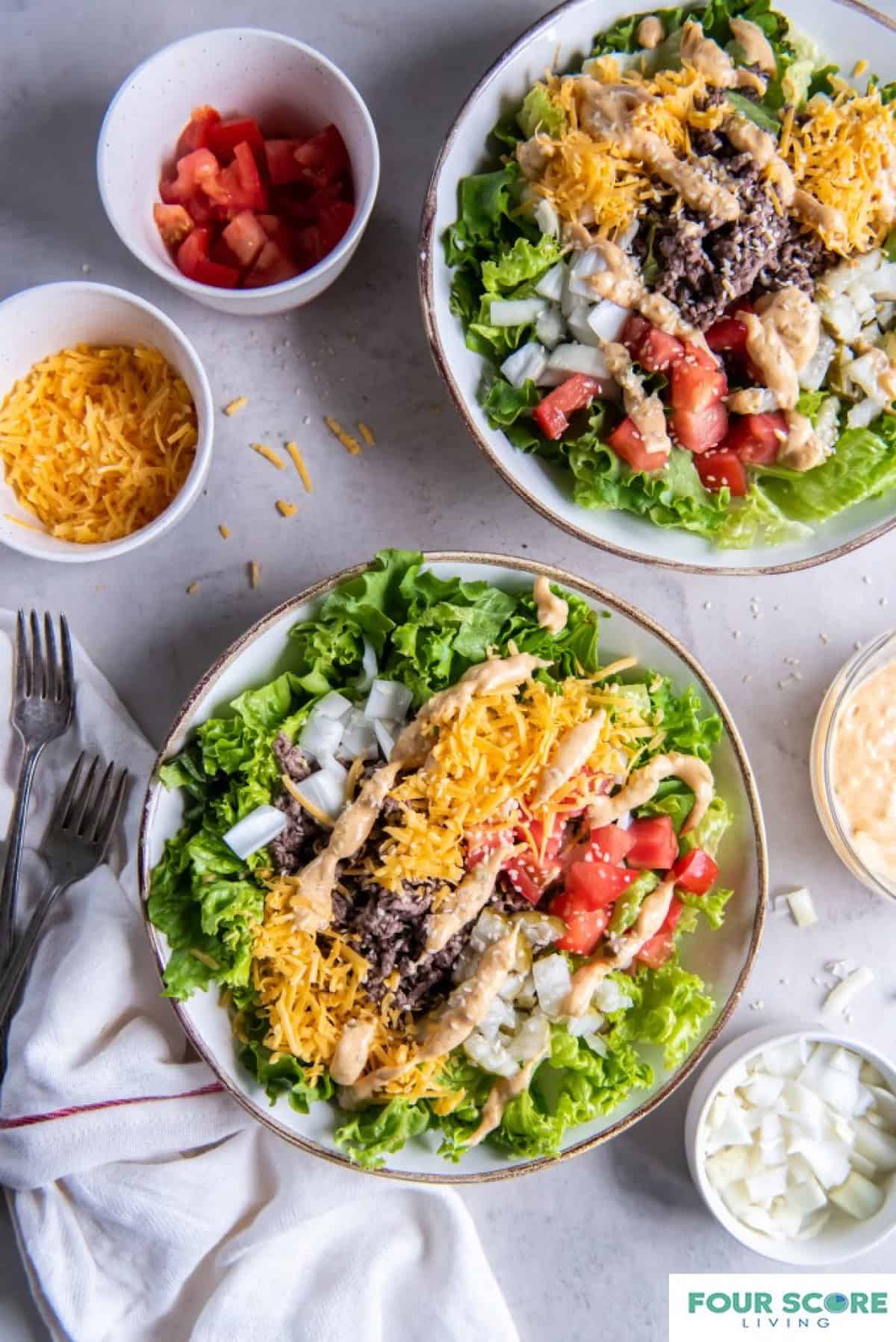 Two plates of salad greens containing cooked ground beef, shredded cheddar cheese, diced red tomatoes, diced white onions, drizzled big mac sauce and a sprinkle of sesame seeds.