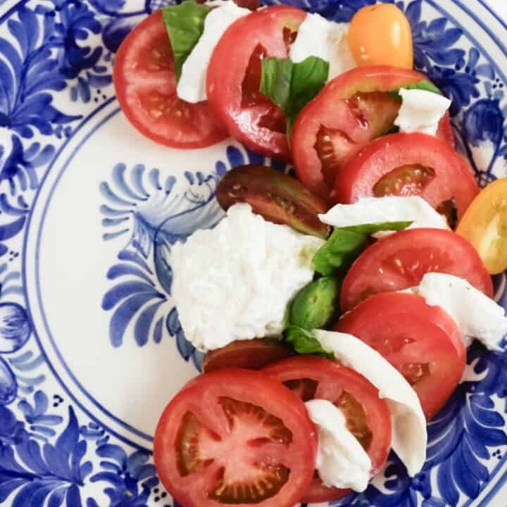Blue and white plate half filled with layers of sliced tomatoes, burrata and basil leaves.