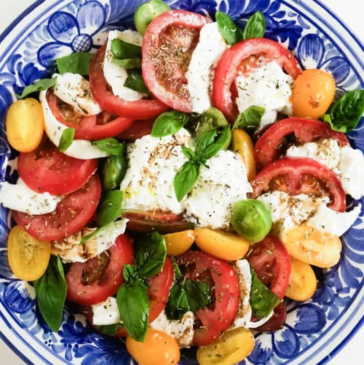 A plate with layers of tomatoes and burrata cheese, topped with basil leaves, dried herbs, olive oil and balsamic vinegar.