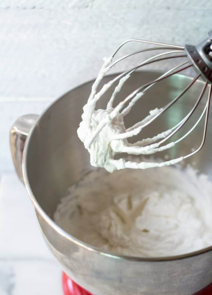 Whipped cream on a stand mixer beater above a bowl of whipped cream.