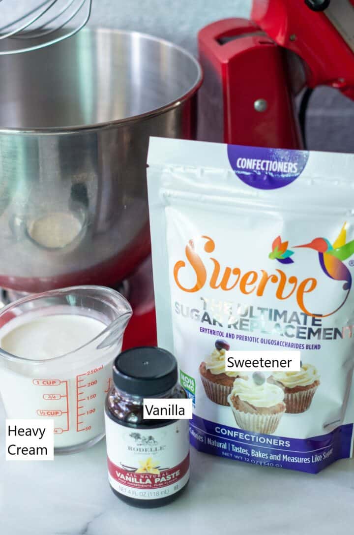 Glass measuring cup filled with heavy cream, a bottle of vanilla paste and a bag of swerve sweetener with a stand mixer behind them.