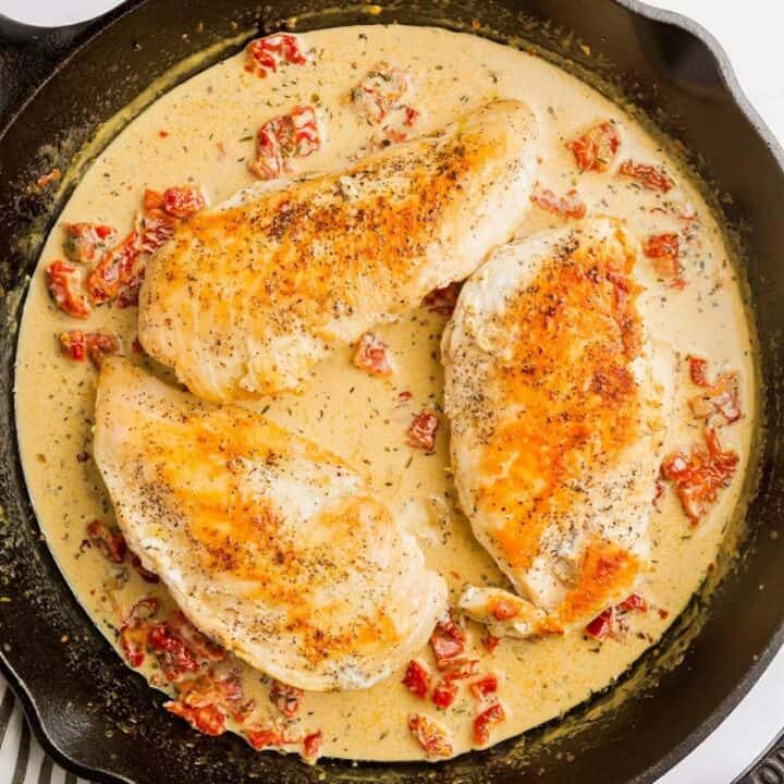 Skillet with cooked chicken breasts being added to herb, sundried tomato cream sauce.