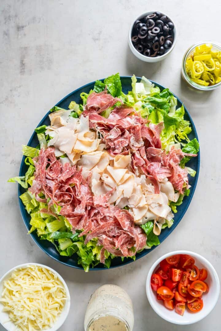 Plate of lettuce topped with sliced deli meats, surrounded by remaining salad ingredients.