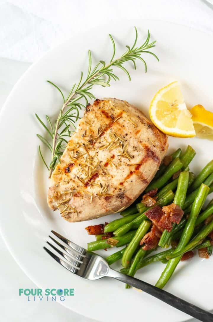 Grilled pork chop on a plate served with green beans, lemon wedges, and a rosemary garnish.