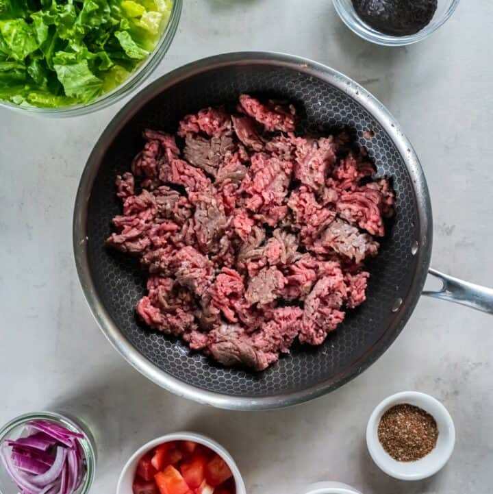 Skillet with ground beef surrounded by individual bowls of taco salad ingredients.