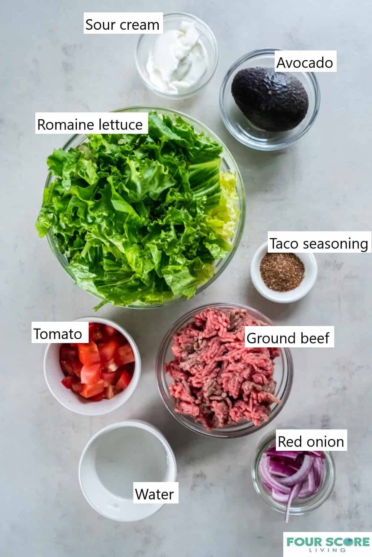 Individual bowls of the ingredients to make a taco salad with chopped lettuce, raw ground beef, chopped tomatoes, taco seasoning, sour cream and a whole avocado.