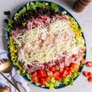 A fully prepared grinder salad on a plate, all ingredients layered including romaine lettuce, red cherry tomatoes, shredded cheese, sliced turkey, salami and capicola.