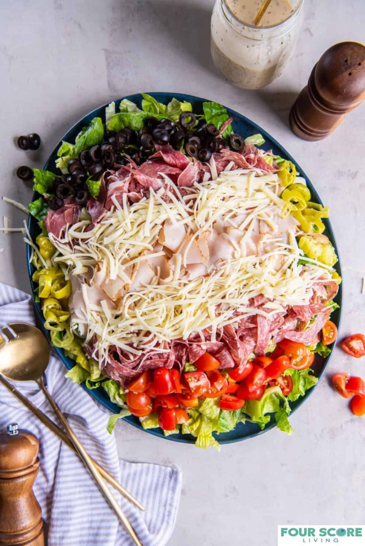 A fully prepared grinder salad on a plate, all ingredients layered including romaine lettuce, red cherry tomatoes, shredded cheese, sliced turkey, salami and capicola.