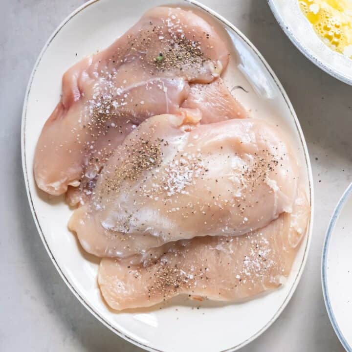 Raw chicken cutlets on a plate seasoned with salt and pepper.