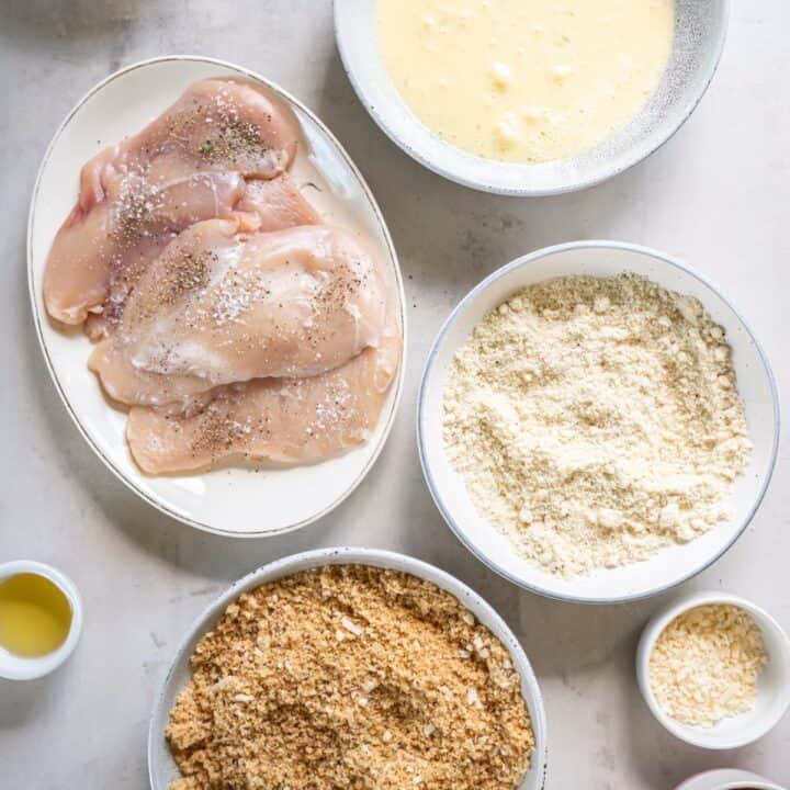 3 shallow bowls with the breading ingredients next to a plate of seasoned chicken cutlets.