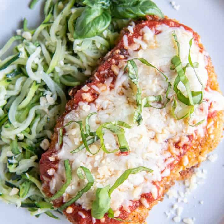 A baked chicken parmesan on a plate with zucchini noodles and topped with chopped basil.