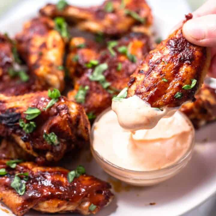 A plate of cooked chicken wings, with one wing being dipped into a dipping sauce.