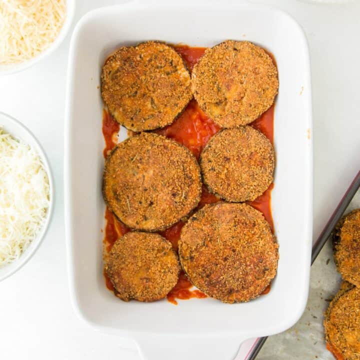 Casserole dish with 6 baked eggplant rounds on a layer of marinara sauce.