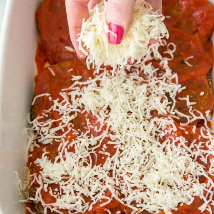 Casserole dish with shredded mozzarella cheese being sprinkled on to eggplant rounds covered in marinara sauce.