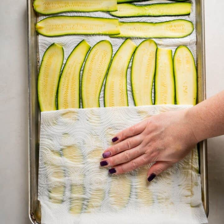 Baking sheet with thin slices of zucchini being dried with a paper towel pressed down by a feminine hand.