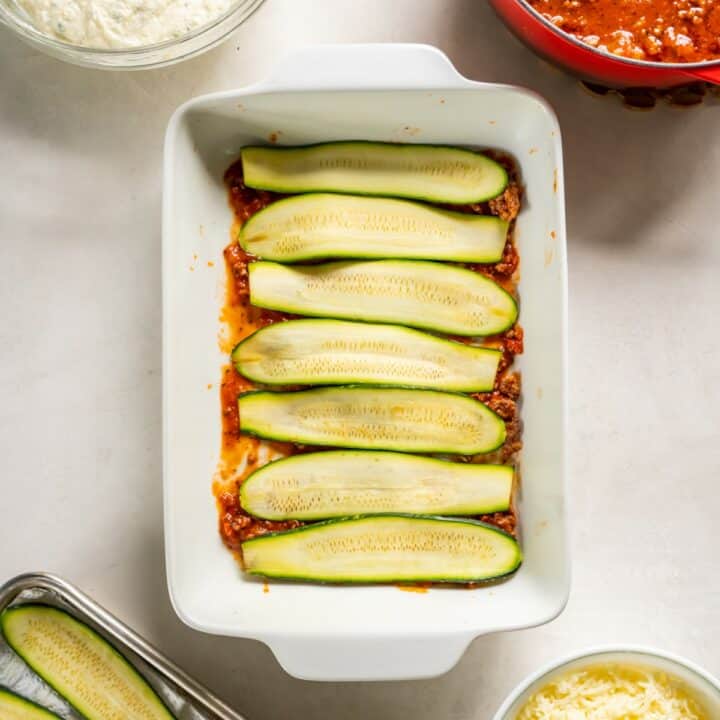 Baking dish with a layer of zucchini being placed on top of the meat sauce.