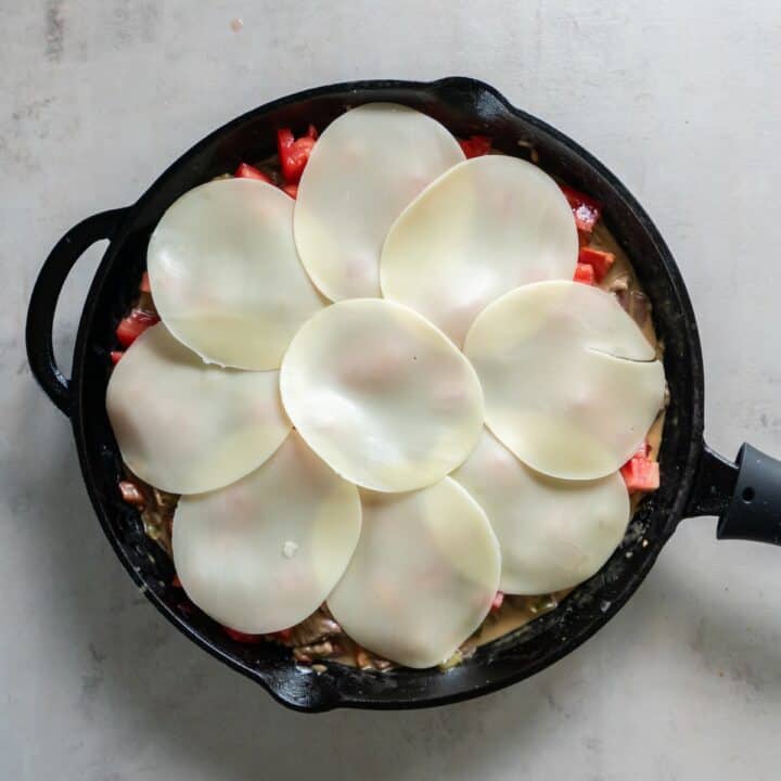 A layer of provolone cheese on top of the cheesesteak ingredients in a cast iron skillet ready to be baked.