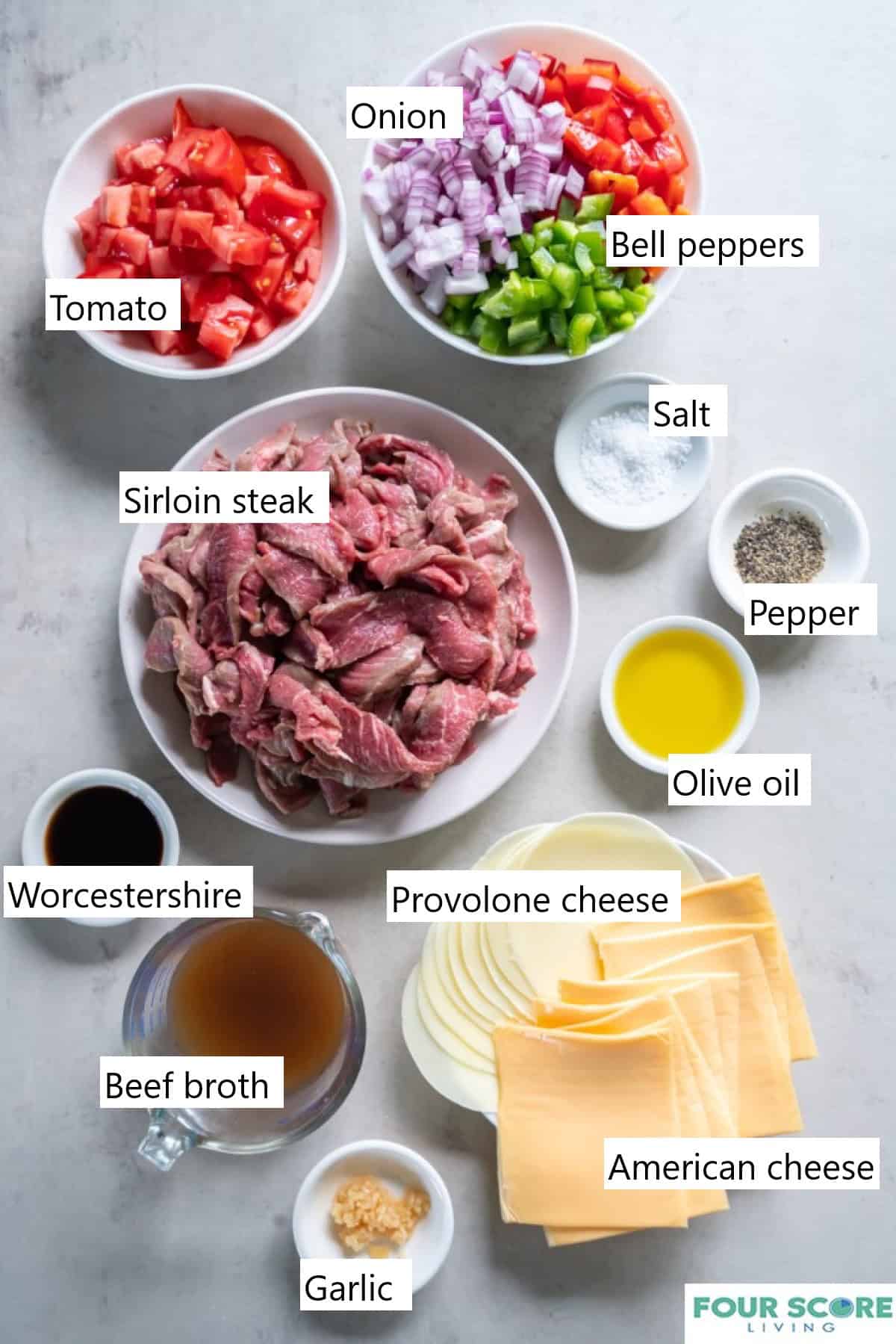 Ingredients to make Philly cheesesteak casserole set out in different sized bowls and dishes, including olive oil, minced garlic, tomatoes, sliced raw steak and chopped peppers and onions.