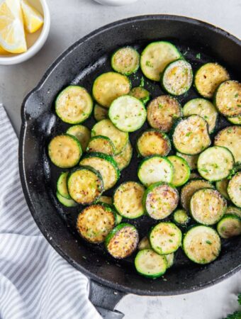 Rounds of sauteed zucchini in a cast iron pan with white bowls of lemon wedges and grated parmesan cheese.