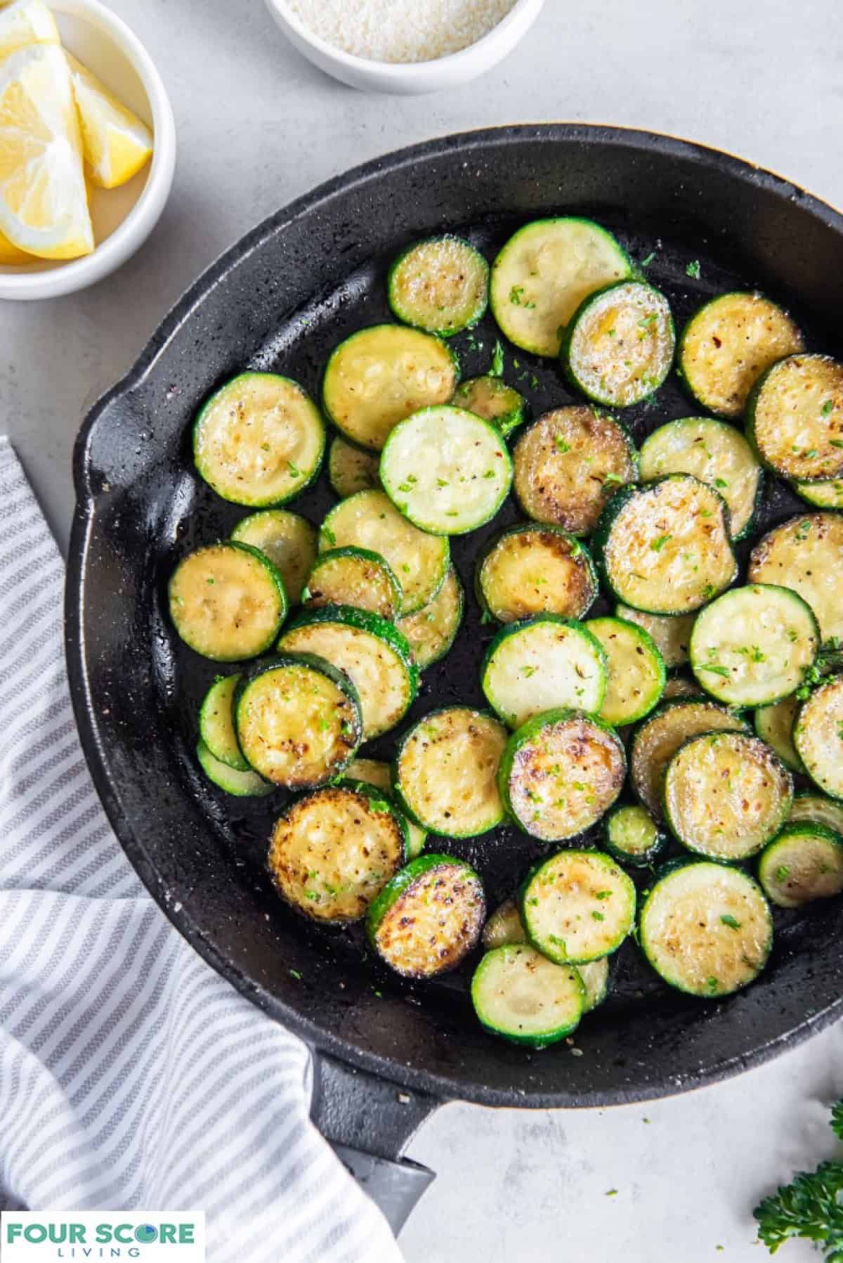 Rounds of sauteed zucchini in a cast iron pan with white bowls of lemon wedges and grated parmesan cheese.