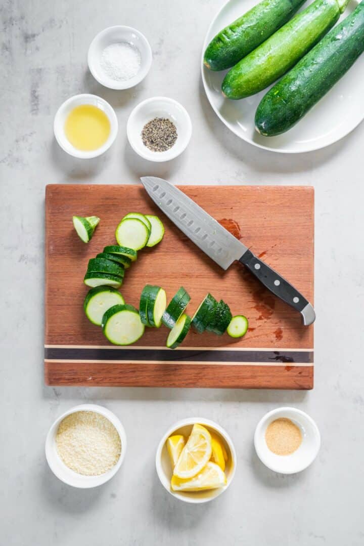 A cutting board with a zucchini being sliced into rounds surrounded by bowls of the remaining ingredients.