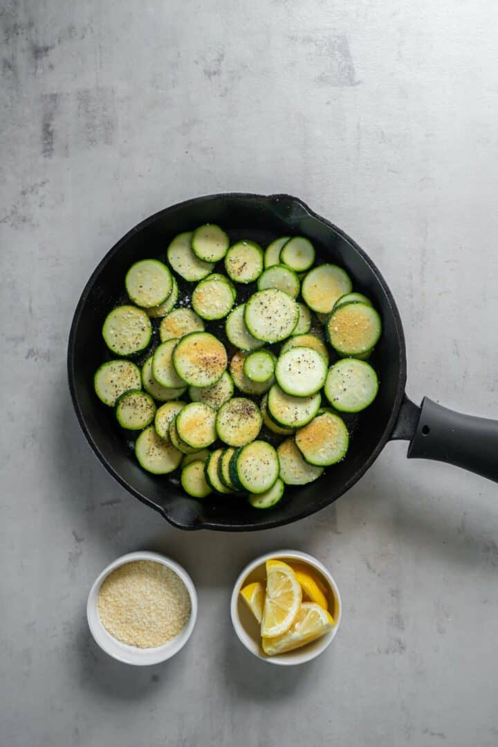 Cast iron skillet with seasoned zucchini rounds. There are also two bowls with lemon wedges and parmesan cheese.