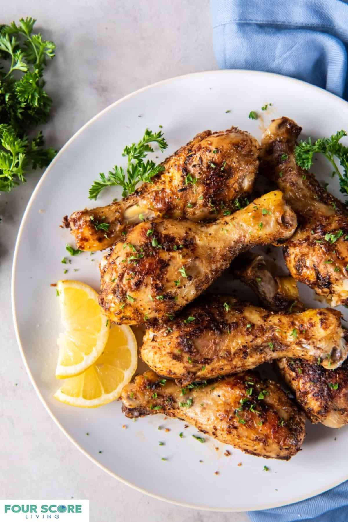 Baked chicken legs plated on a white plate with lemon slices and chopped fresh herb garnish.