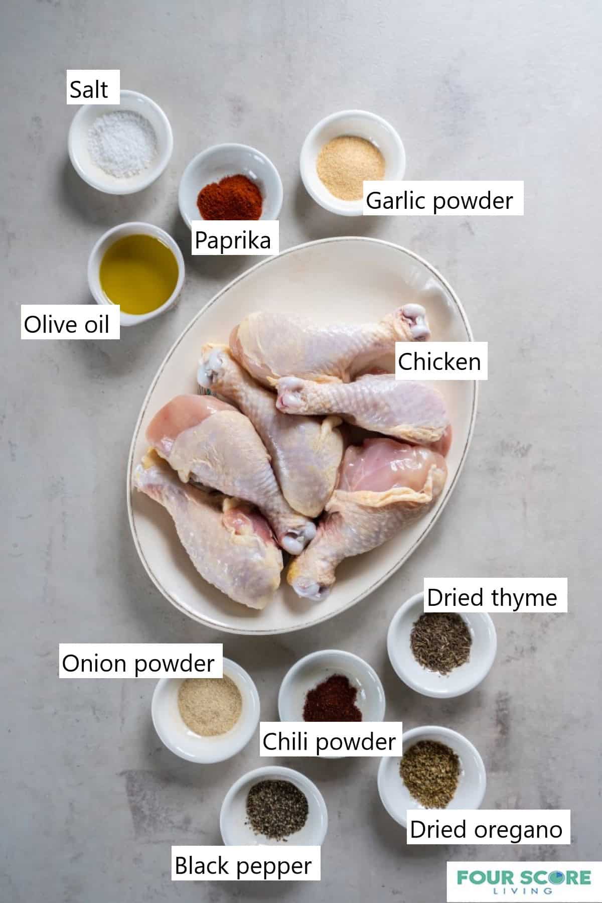 Ingredients to make baked chicken legs, including 6 uncooked chicken legs on an oval white platter and small white dishes of herbs and spices.