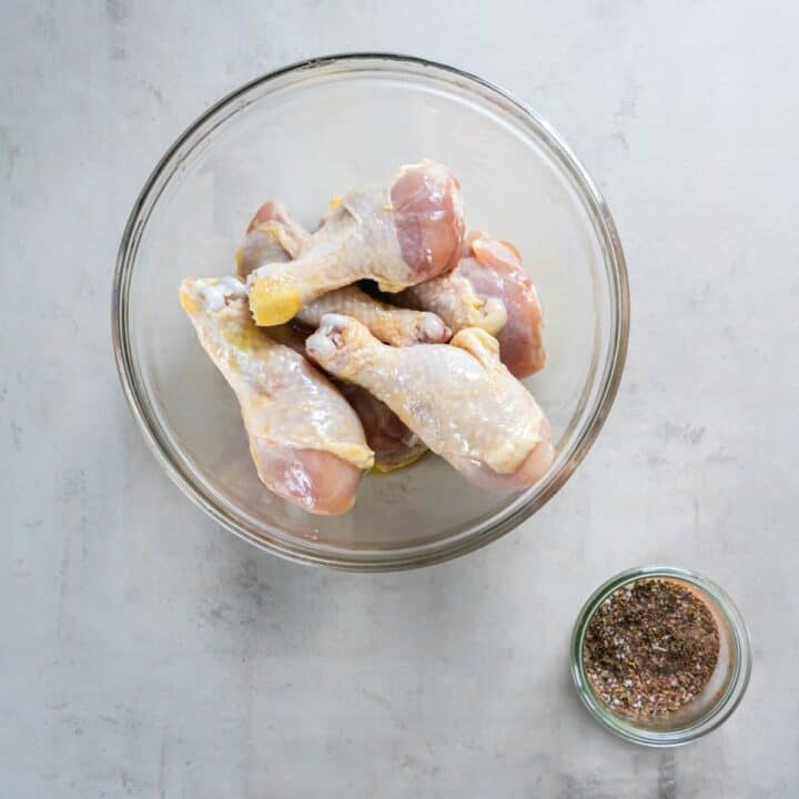 Glass bowl of oiled chicken legs and a small glass bowl of mixed seasoning blend.