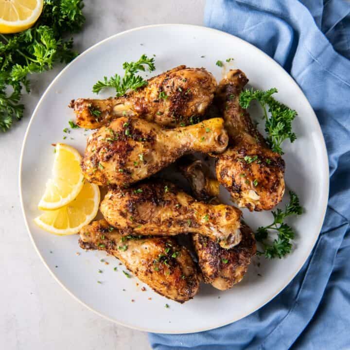 Plate with six baked chicken legs, garnished with chopped parsley and lemon wedges.