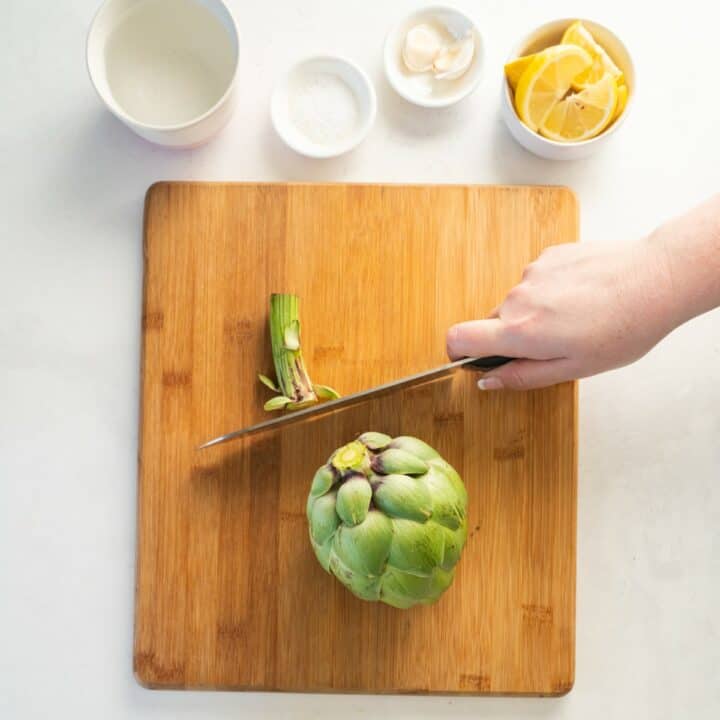 Stem being cut off of an artichoke on a wooden cutting board.