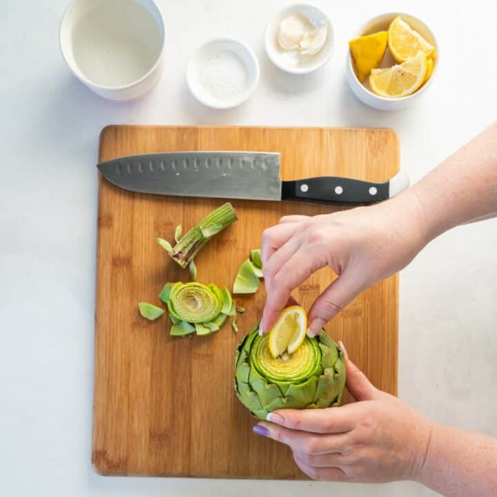 A lemon wedge is being squeezed onto the cut surface of an artichoke.