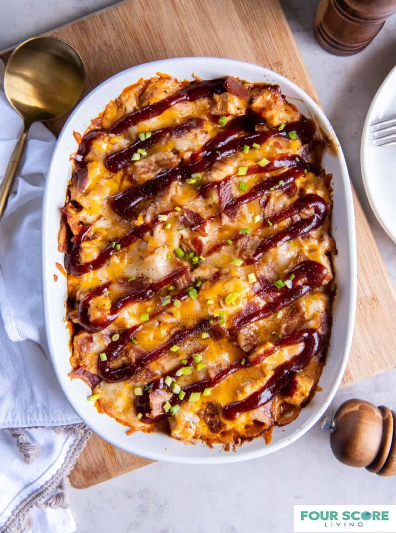 Oval casserole dish with a baked BBQ chicken casserole drizzled with BBQ sauce and set on a wooden cutting board next to a kitchen towel and a large round serving spoon.