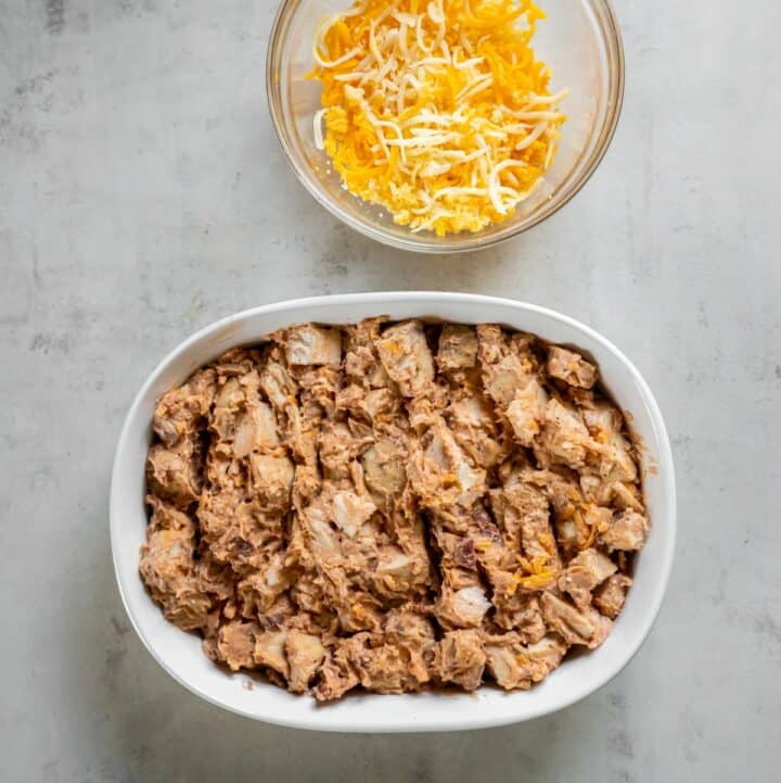 Casserole dish containing the cheesy BBQ chicken mixture with a bowl of shredded cheese next to it.