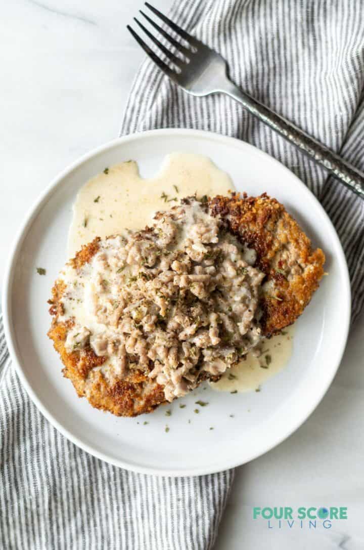 Chicken fried steak with sausage gravy on a round plate next to a fork and napkin.