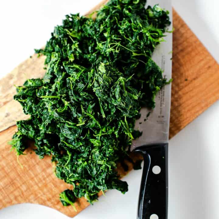 Thawed frozen spinach being chopped fine on a wooden cutting board.