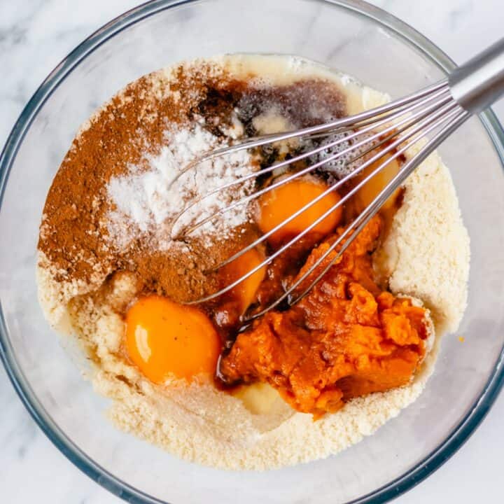 Glass bowl with the wet ingredients, eggs, oil, syrup, pumpkin puree and vanilla extract being added to the flour mixture.