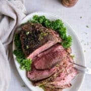 A cooked sirloin tip roast on an oval shaped plate. Half of the sirloin tip is sliced into slices and a two tong serving tool is piercing the slices. There is a green garnish on the plate and wooden salt and pepper shakers in the background.