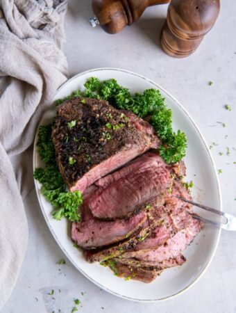 A cooked sirloin tip roast on an oval shaped plate. Half of the sirloin tip is sliced into slices and a two tong serving tool is piercing the slices. There is a green garnish on the plate and wooden salt and pepper shakers in the background.