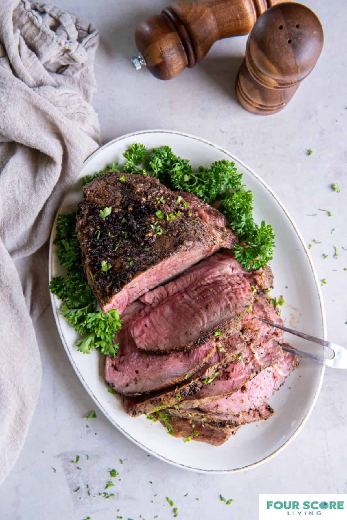 A cooked sirloin tip roast on an oval shaped plate. Half of the sirloin tip is sliced into slices and a two tong serving tool is piercing the slices. There is a green garnish on the plate and wooden salt and pepper shakers in the background.