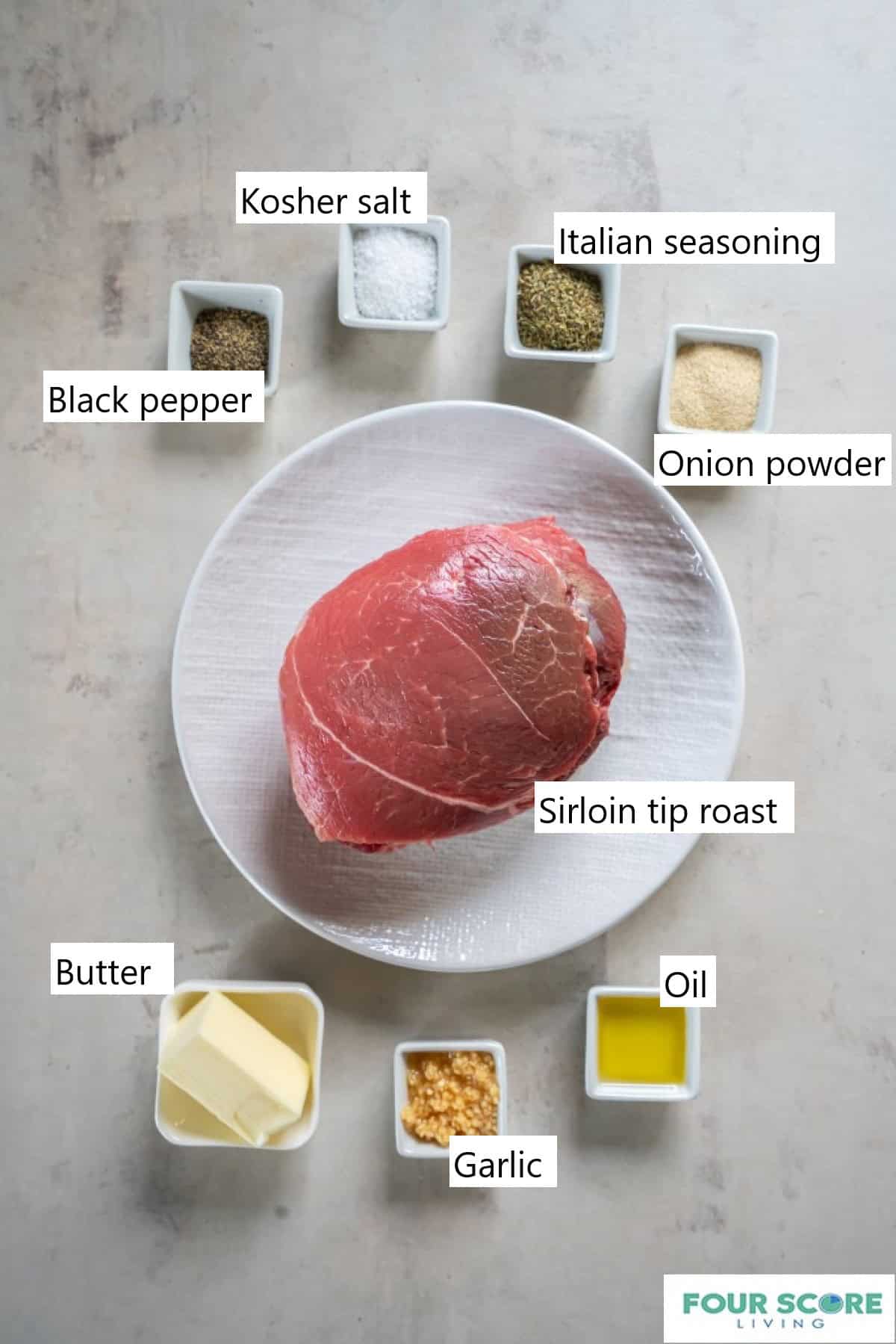 A raw sirloin tip roast placed on a round plate. Surrounded by small dishes with additional ingredients: butter, oil, minced garlic, salt, spices.