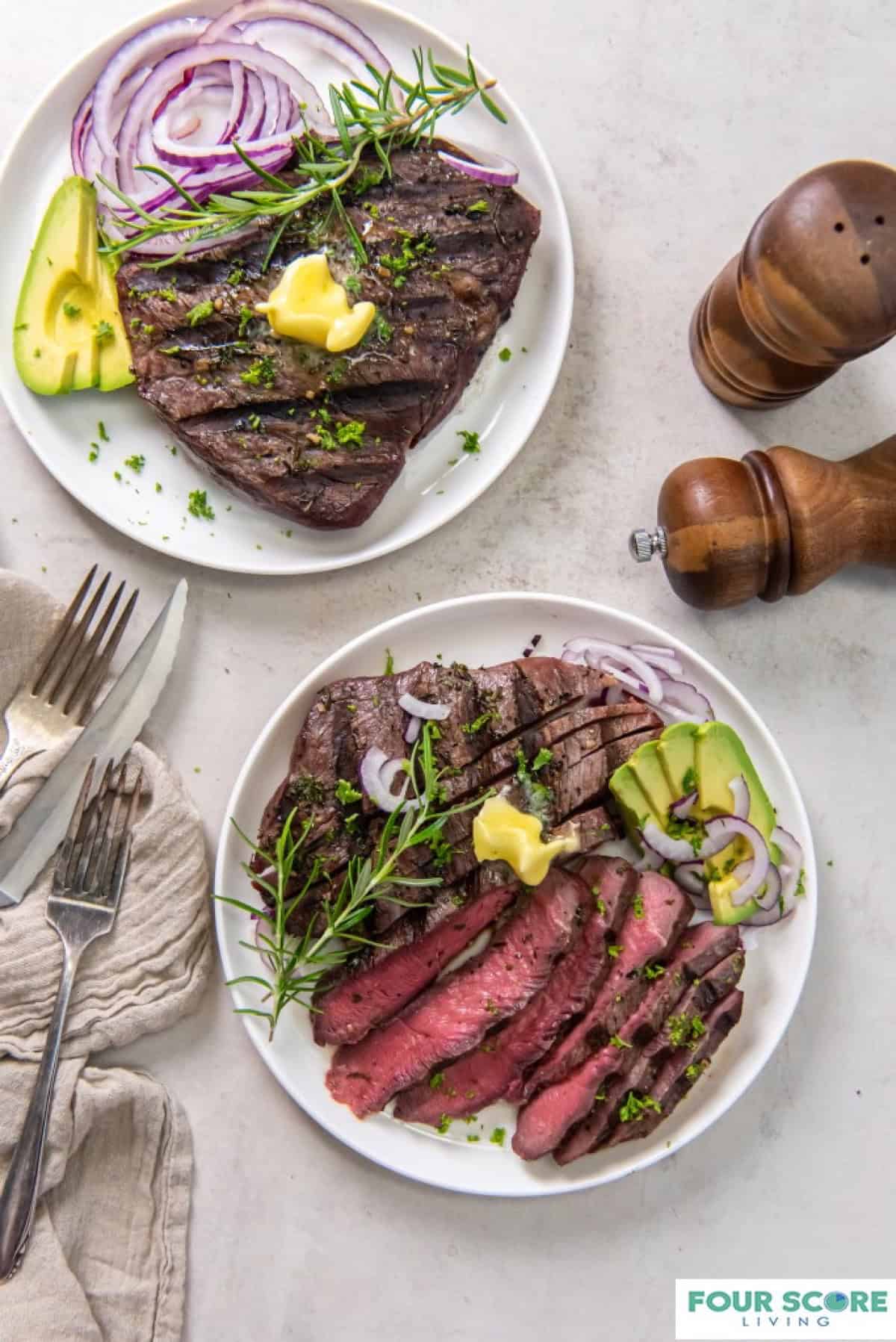 Two white plates of sirloin tip steak, one sliced and each garnished with sliced avocado, purple onion and a pat of butter.