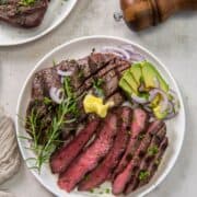 Two white plates of sirloin tip steak, one sliced and each garnished with sliced avocado, purple onion and a pat of butter.