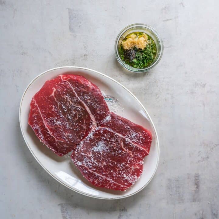 Plate with salted steaks and a small glass bowl with the fresh herbs, oil, garlic and pepper being mixed together.