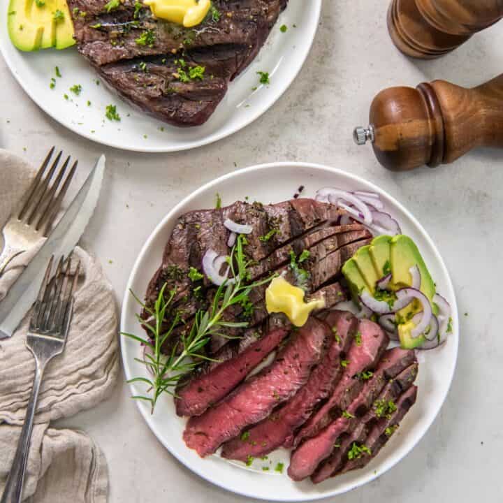 A plate with a grilled sirloin tip steak sliced and garnished with sliced avocado, purple onion and a pat of butter.