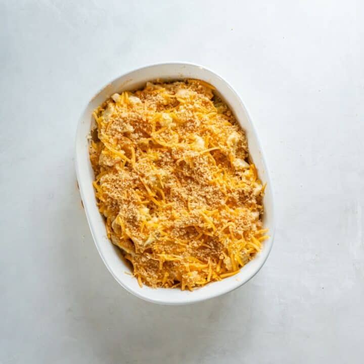 Baking dish filled with unbaked keto cauliflower mac and cheese, topped with shredded cheddar cheese and crushed pork rinds.